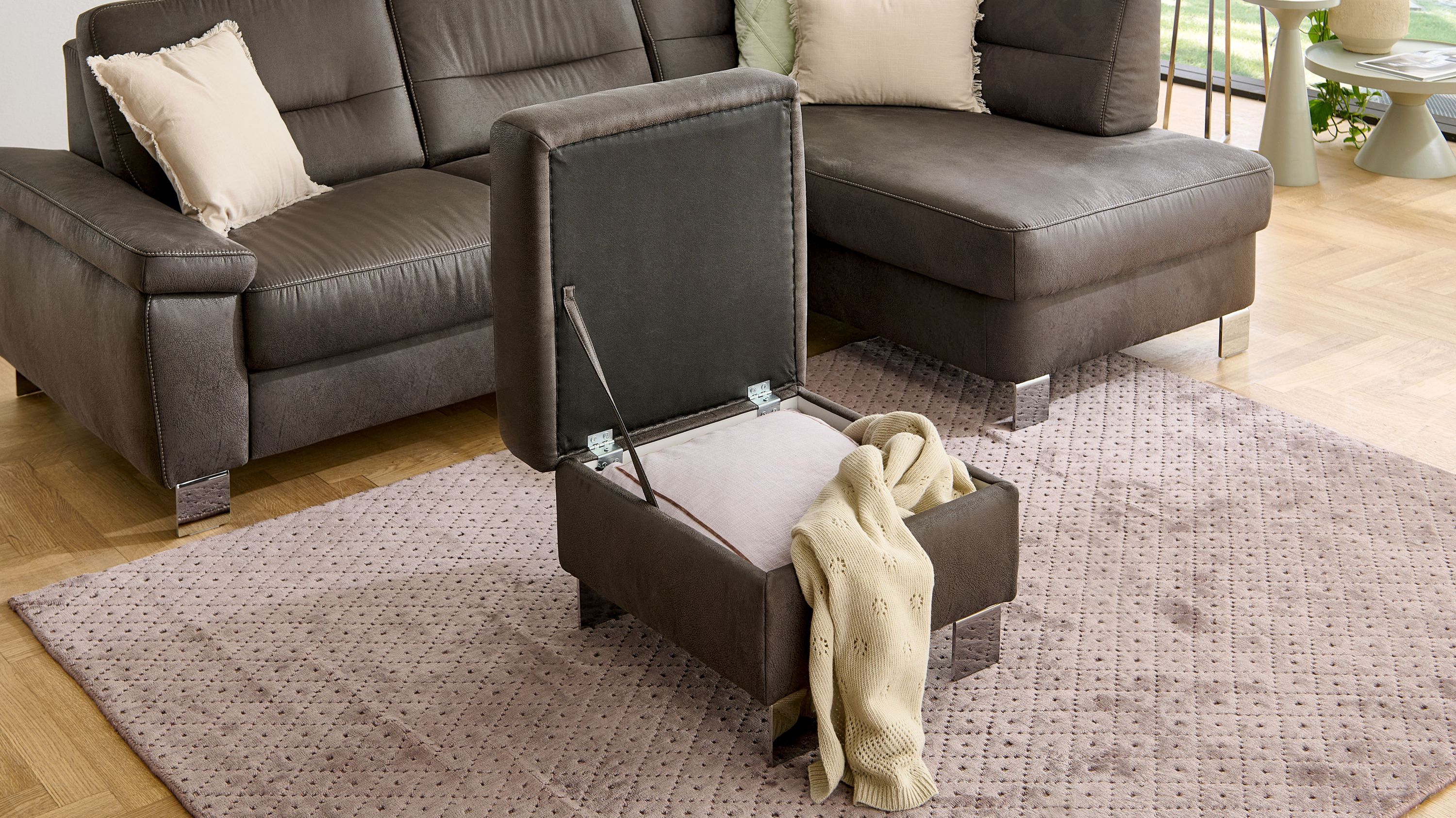 Quadratischer Polsterhocker mit aufklappbarem Sitz und Stauraum in dunkelgrauer Farbe auf Teppich vor Sofa