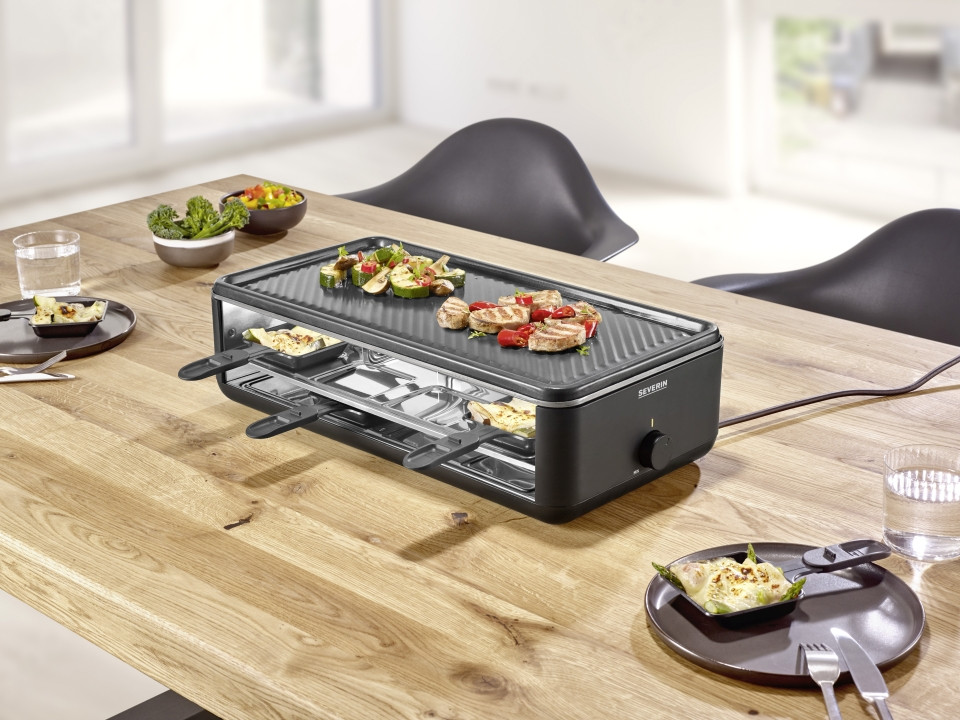 Raclette-Grill mit Keramikplatte und 8 Pfännchen auf gedecktem Holztisch leicht schräg von vorne rechts fotografiert