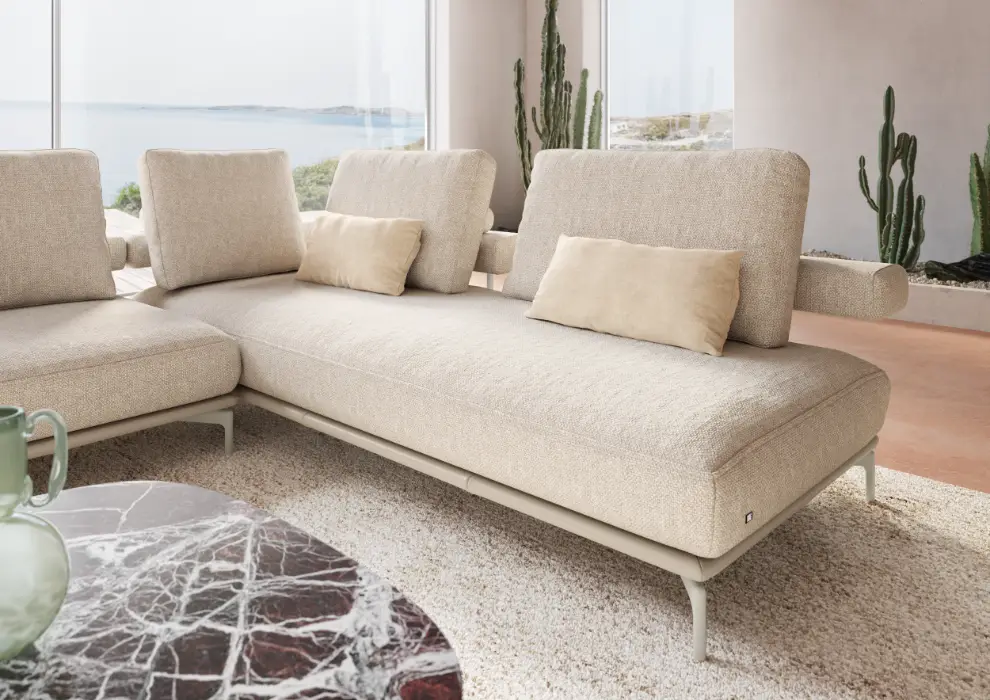 Modernes beiges Sofa mit verstellbaren Rückenlehnen und Zierkissen, platziert in einem hellen Wohnzimmer mit Meerblick und dekorativen Kakteen.