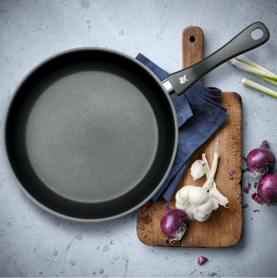 Antihaft-Bratpfanne in Draufsicht auf Küchentisch mit Schneidebrett, Knoblauch, roten Zwiebeln und Frühlingszwiebeln.