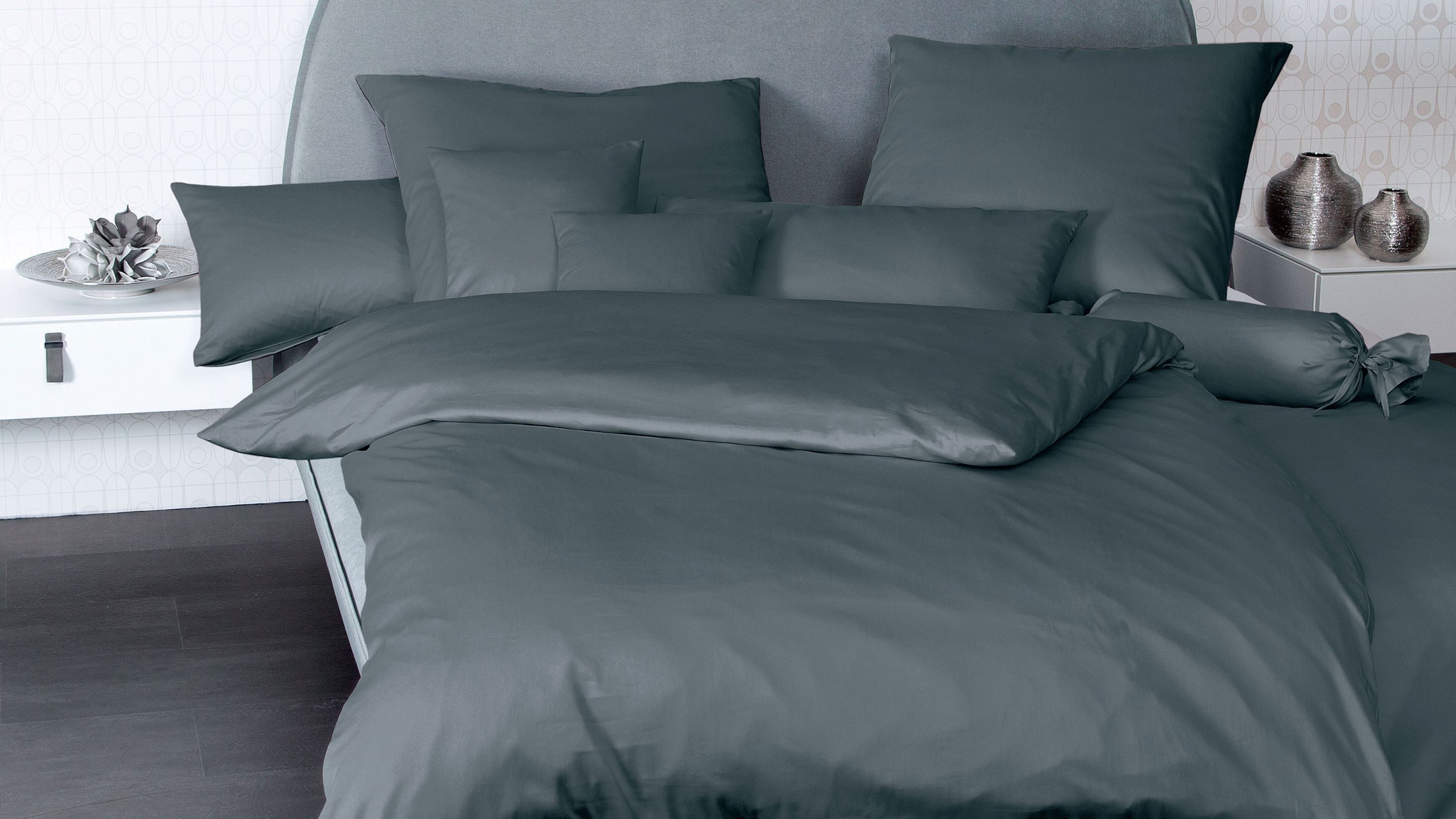 Anthrazitfarbene Bettwäsche mit mehreren Kissen und Zierkissen auf einem Bett mit grauem Kopfteil