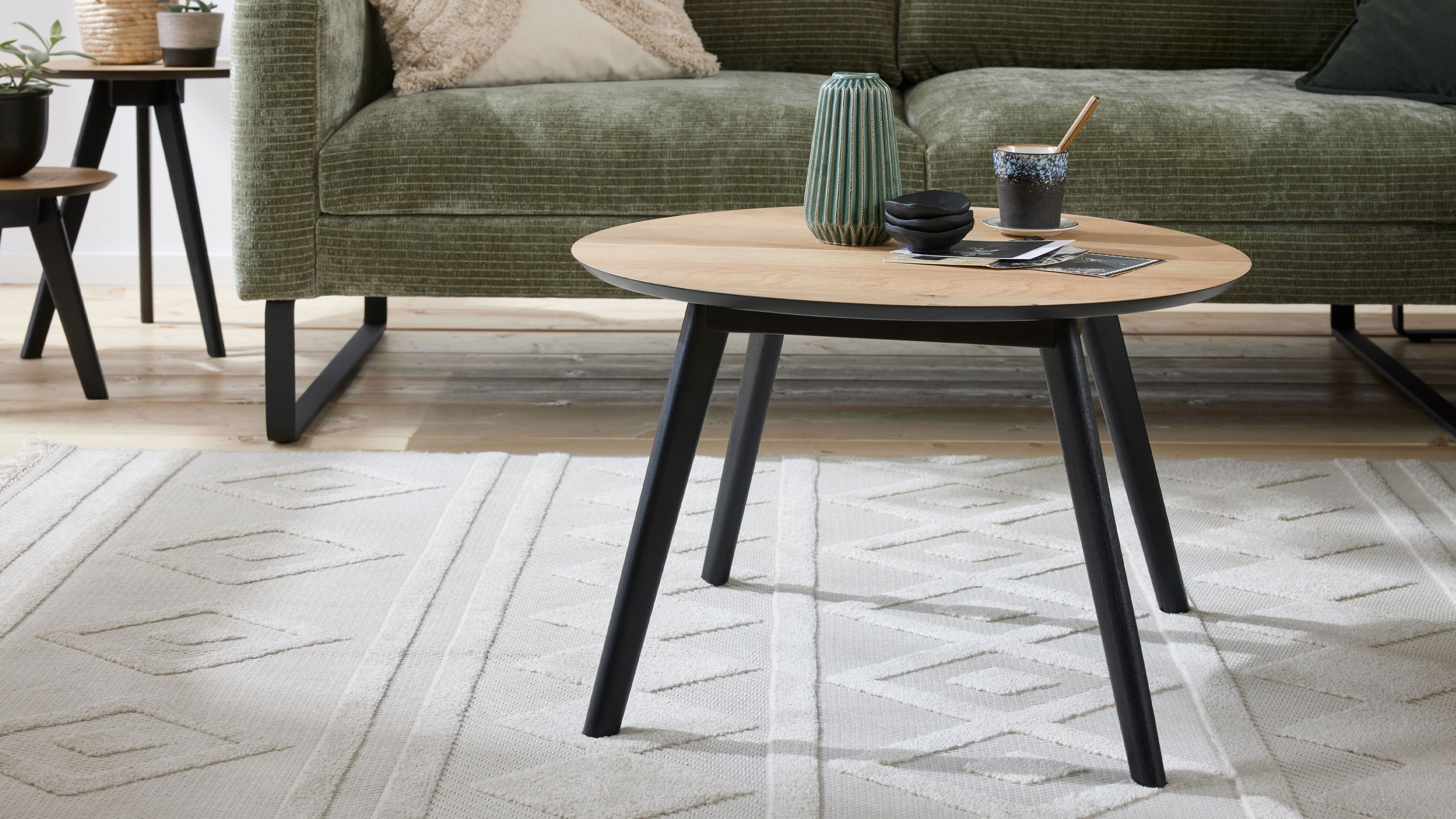 Runder Couchtisch mit hellem Holzfarbton und schwarzen schrägen Beinen auf hellem Teppich vor grünem Sofa