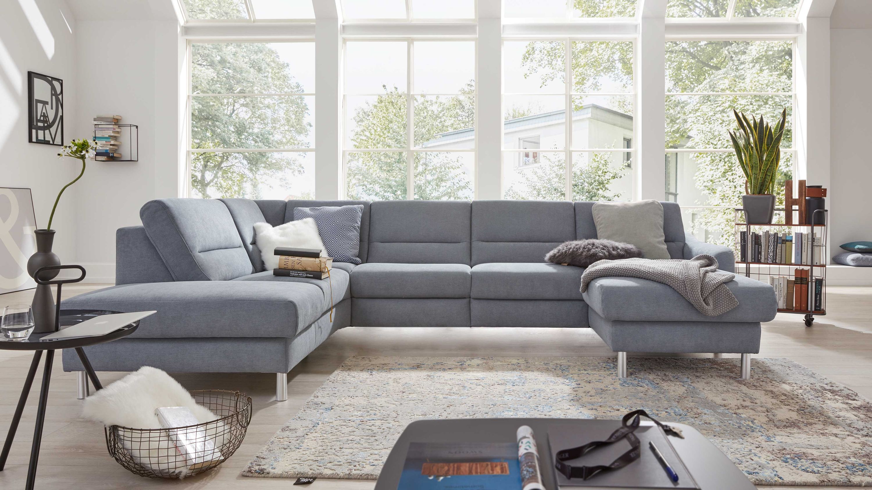 Ecksofa in eisblau mit Metallfüßen und mehreren Kissen in einem hellen Wohnzimmer vor großen Fenstern