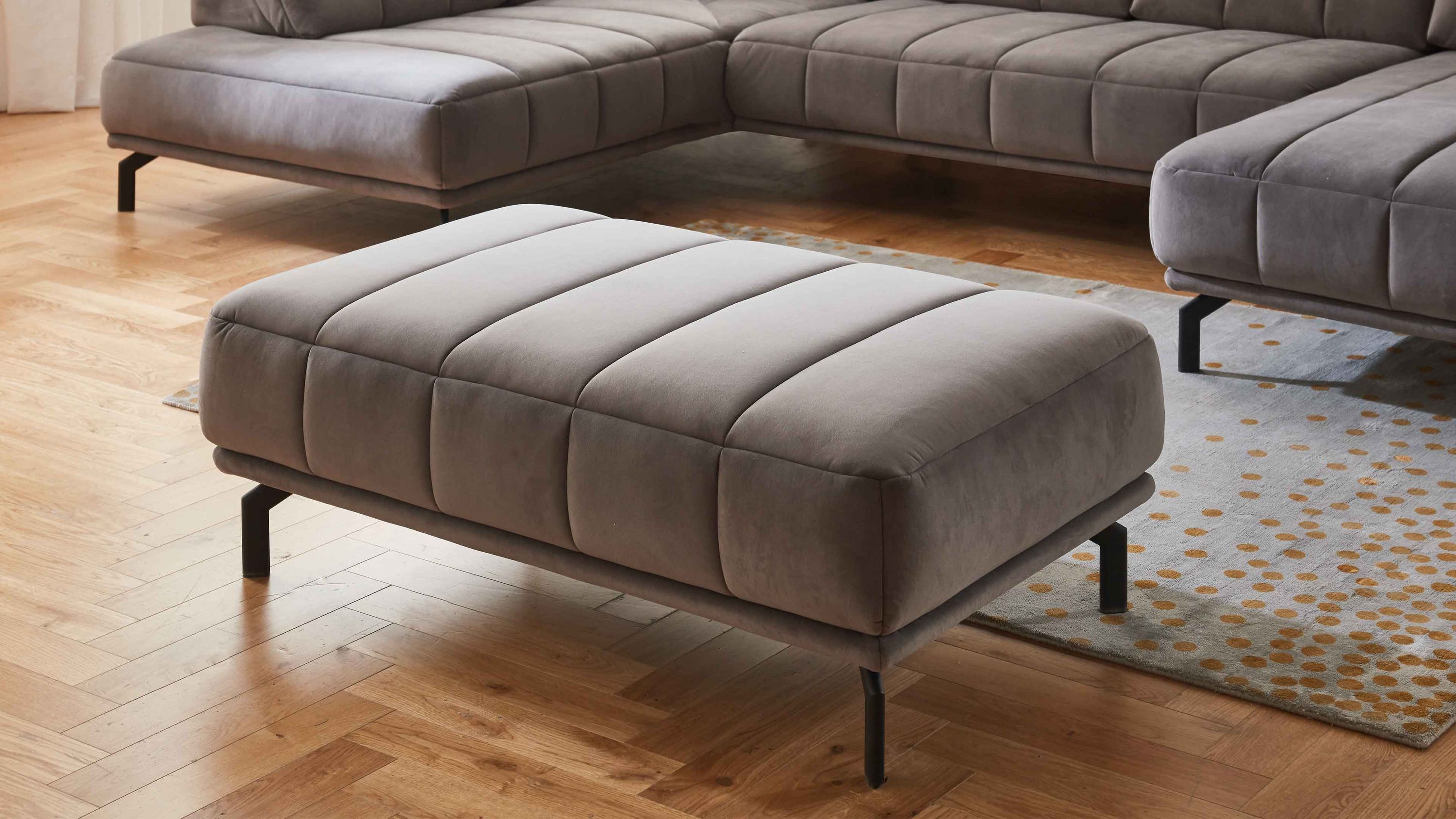 Großer grauer Hocker mit gepolsterter Oberfläche und schwarzen Metallfüßen vor grauem Sofa auf Holzboden
