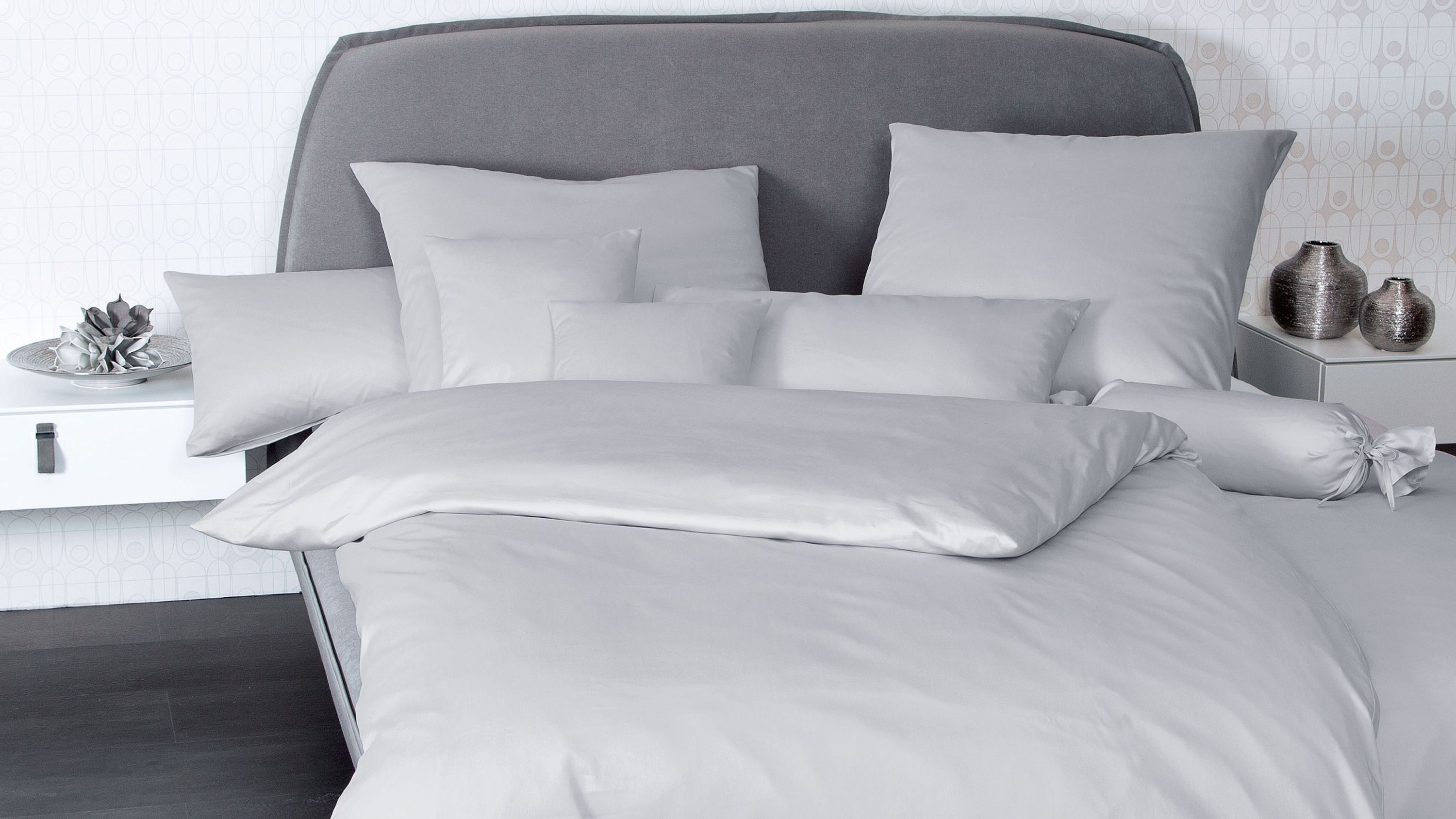 Hellgraue Bettwäsche mit mehreren Kissen und einer Rundrolle auf einem Bett mit grauem Kopfteil