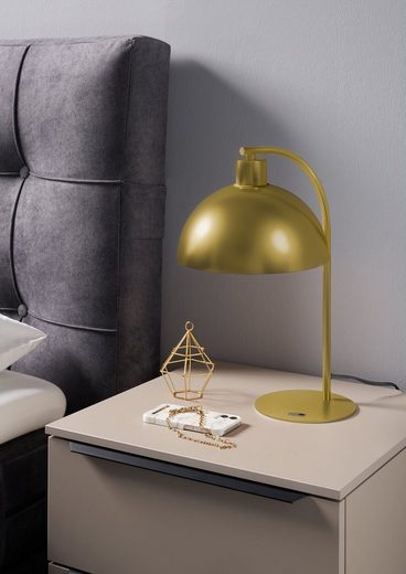 Nachttisch mit goldener Lampe, Buch und Dekoobjekt neben grauem Kopfteil, aus frontaler Perspektive fotografiert.