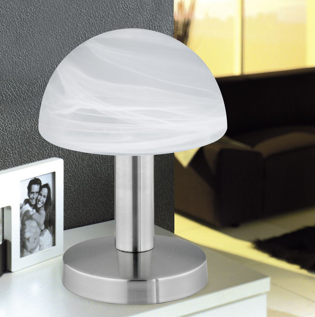 Klassische Tischlampe mit weißem Schirm auf hellem Beistelltisch im Hintergrund Couch und beleuchtetes Regal