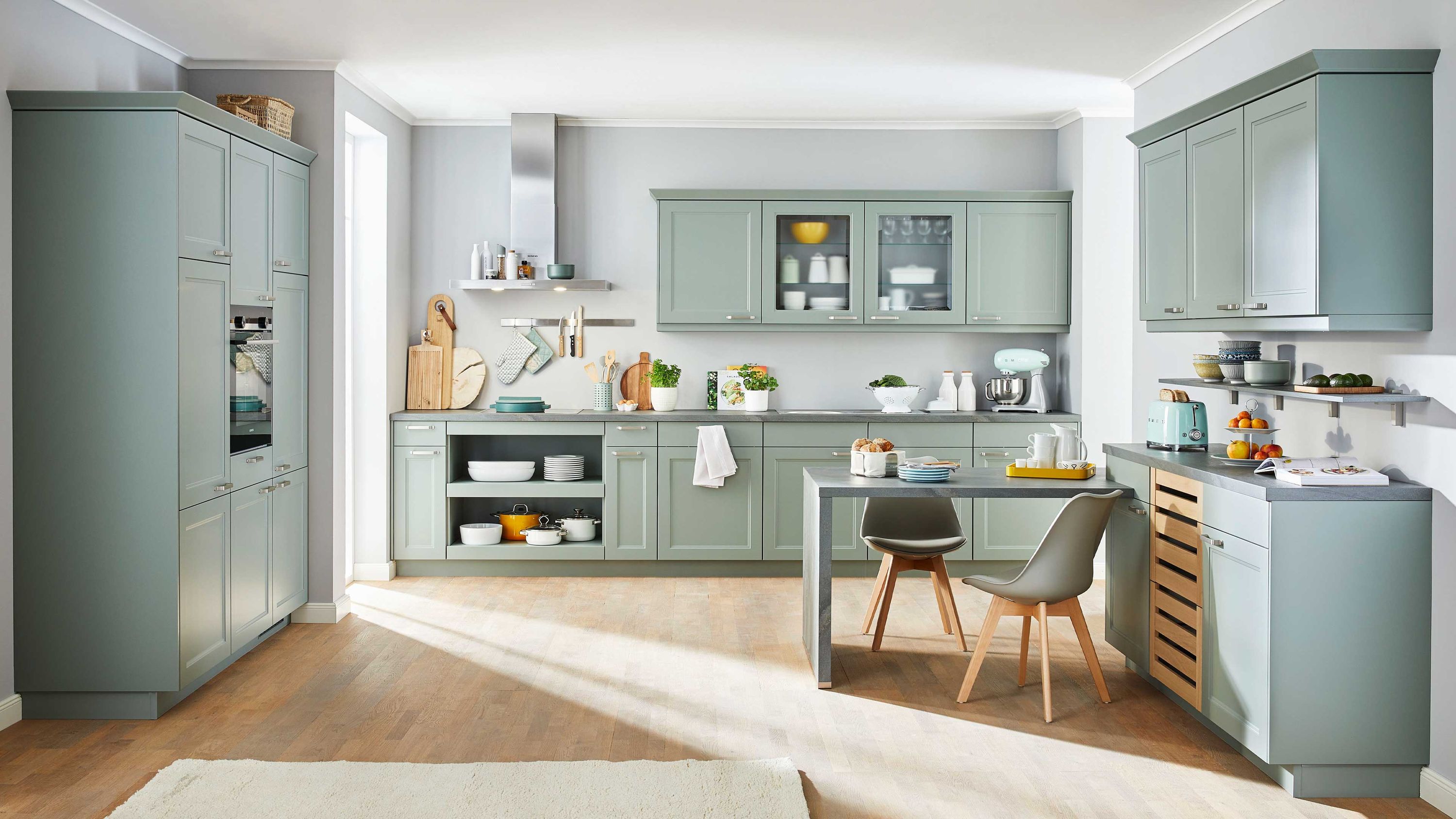 Pastellgrüne Landhaus-Winkelküche mit Glashängeschränken. Pastellgrüne Winkelküche im Landhausstil mit Glashängeschränken offenen Regalen Einbaugeräten und Essbereich.