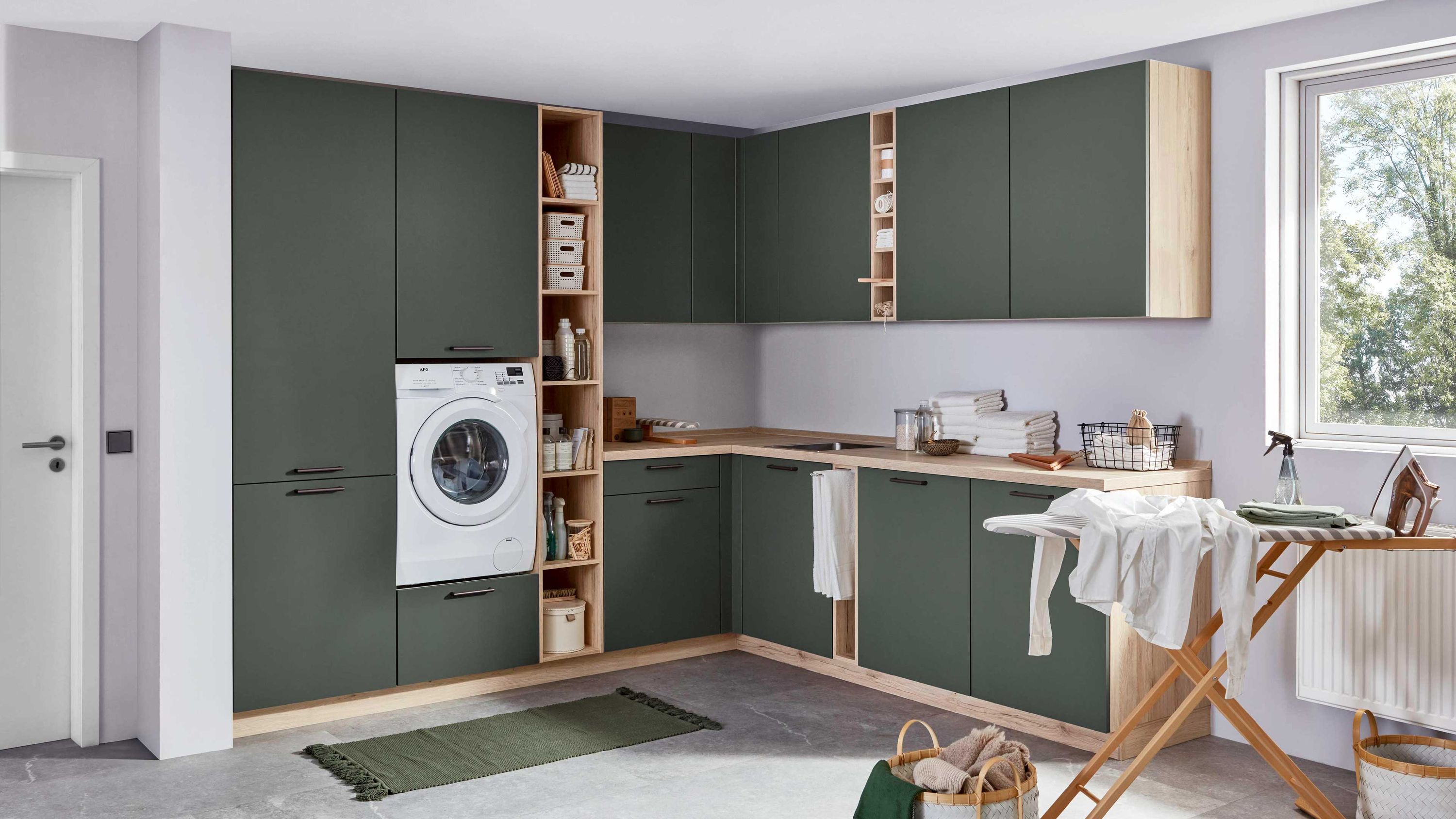 Eckküche in Dunkelgrün mit Holzregalen integrierter Waschmaschine heller Arbeitsplatte und großem Fenster.