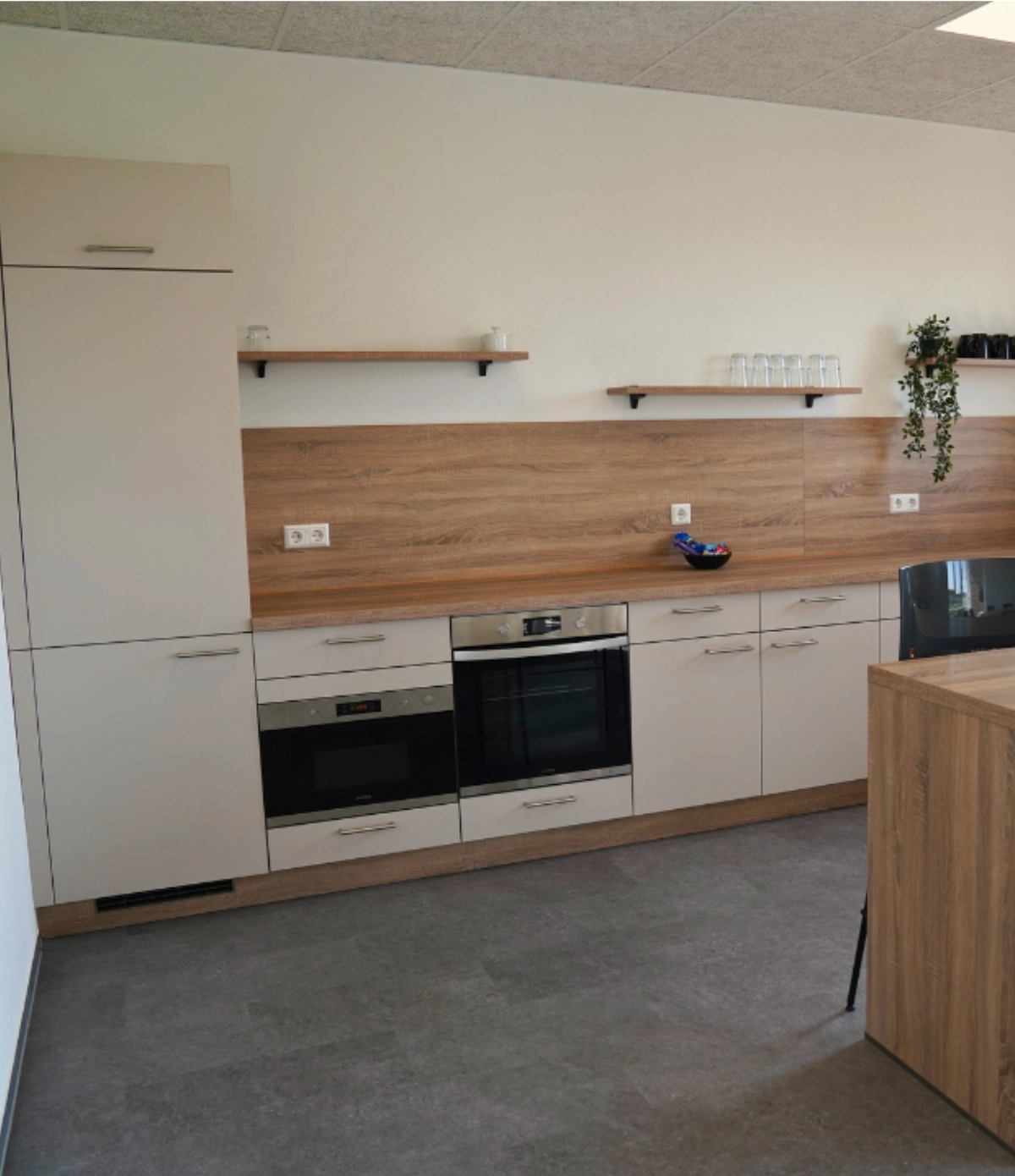 Moderne Büroküche mit Einbaugeräten, Holzarbeitsplatte, Hängeregalen und Stauraum, funktionaler Pausenraum in zeitgemäßem Office‑Design