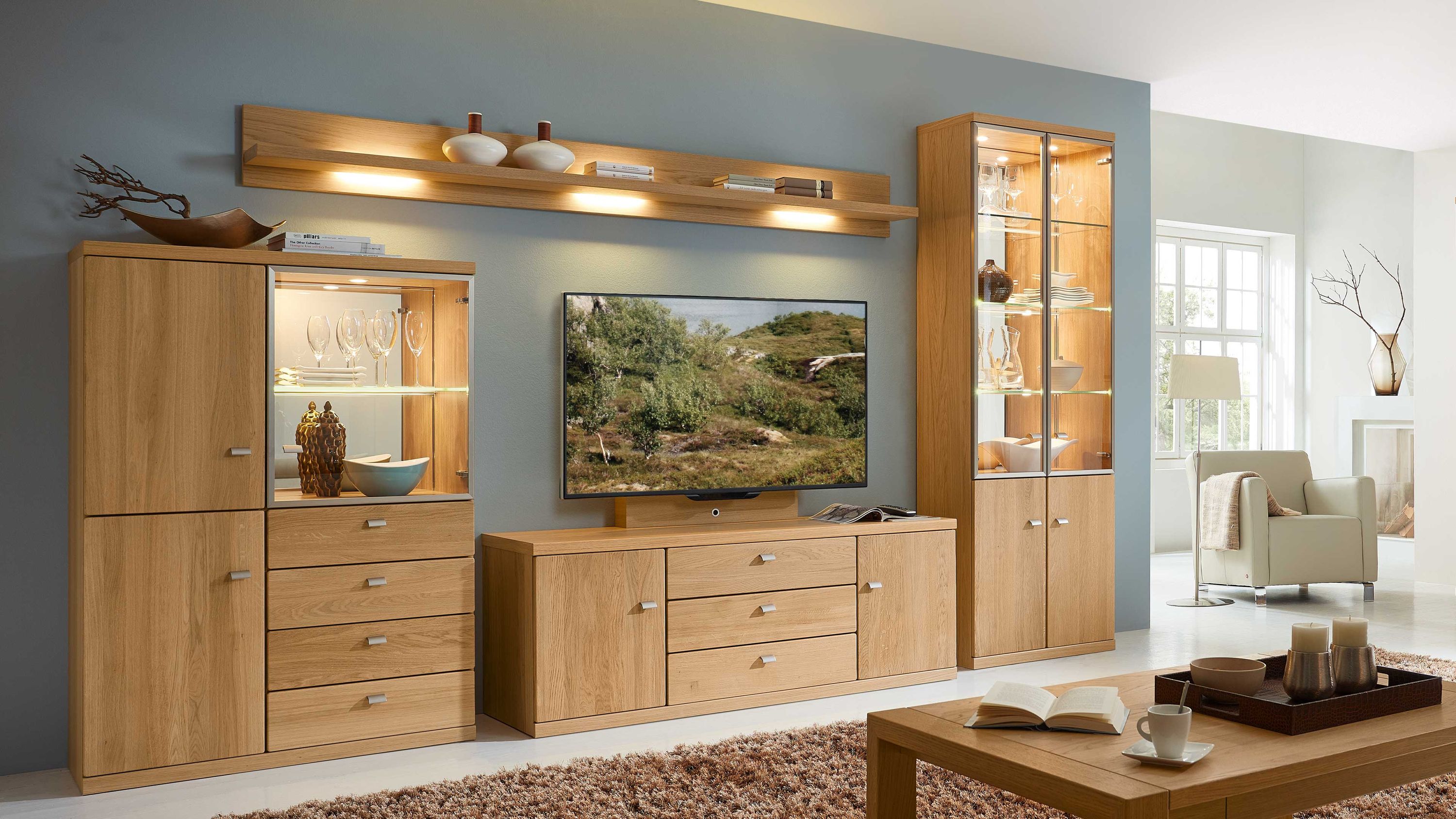 Wohnkombination aus Holz mit Schränken und Wandregal Wohnkombination aus mehreren Schränken und einem Wandregal in hellbrauner Holzoptik mit Glastüren und Schubladen