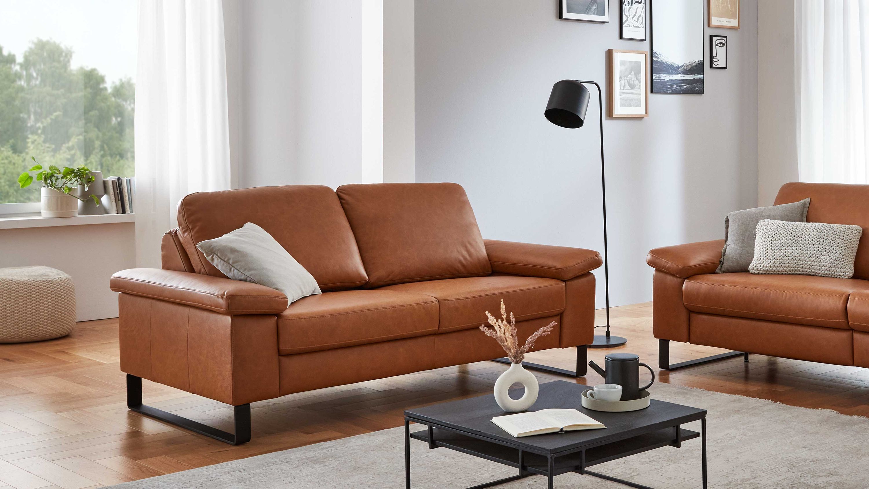 Zweisitziges Sofa aus cognacfarbenem Leder mit schwarzen Metallfüßen und zwei Kissen in einem hellen Wohnzimmer