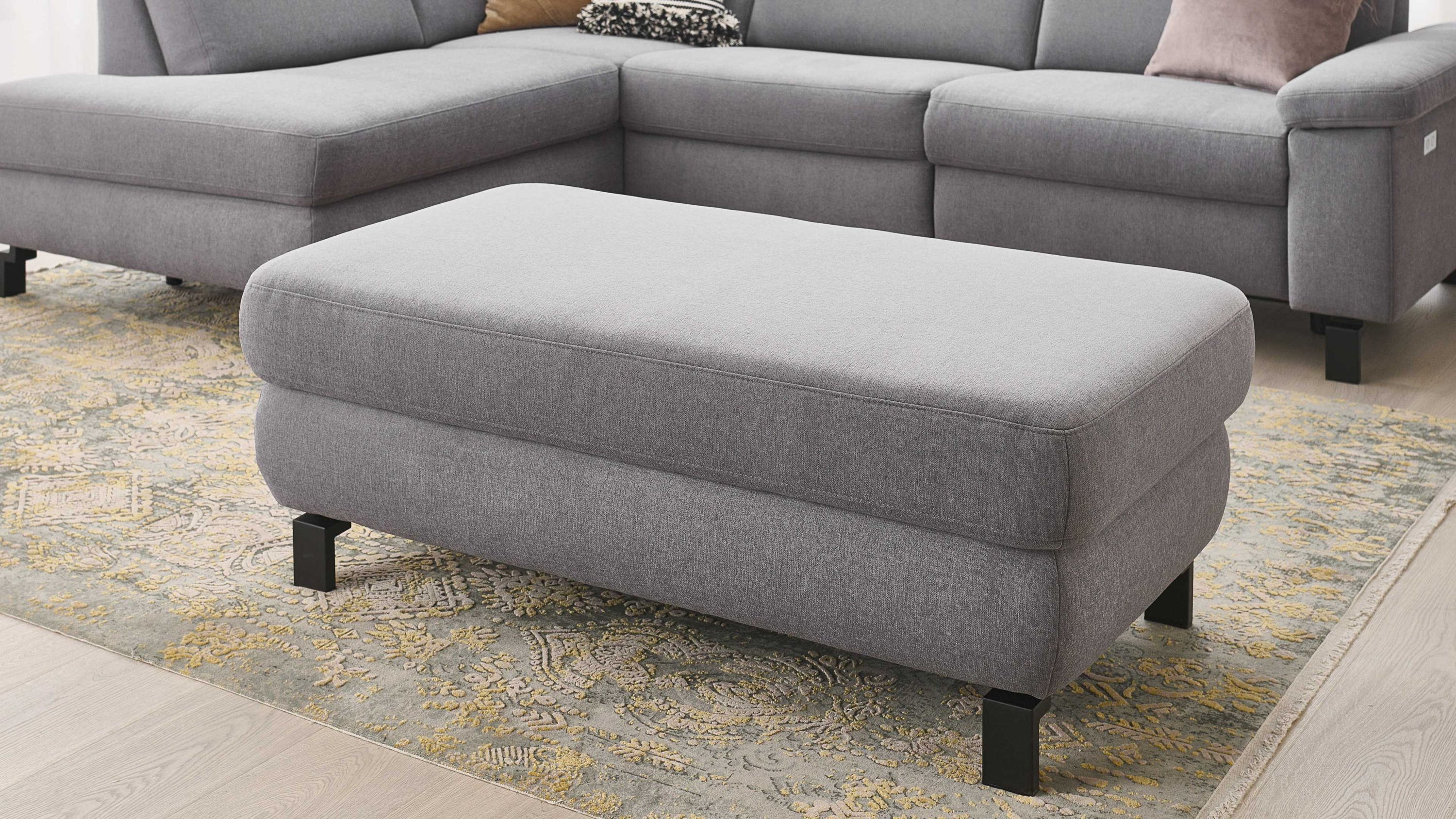 Rechteckiger Hocker mit platingrauem Stoffbezug und schwarzen Metallfüßen vor grauem Ecksofa