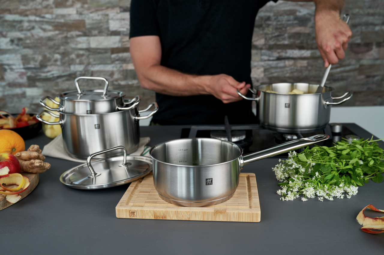 Edelstahl-Kochtöpfe und Stieltopf auf Herd, Person rührt Suppe, frische Kräuter und Zutaten auf der Arbeitsfläche