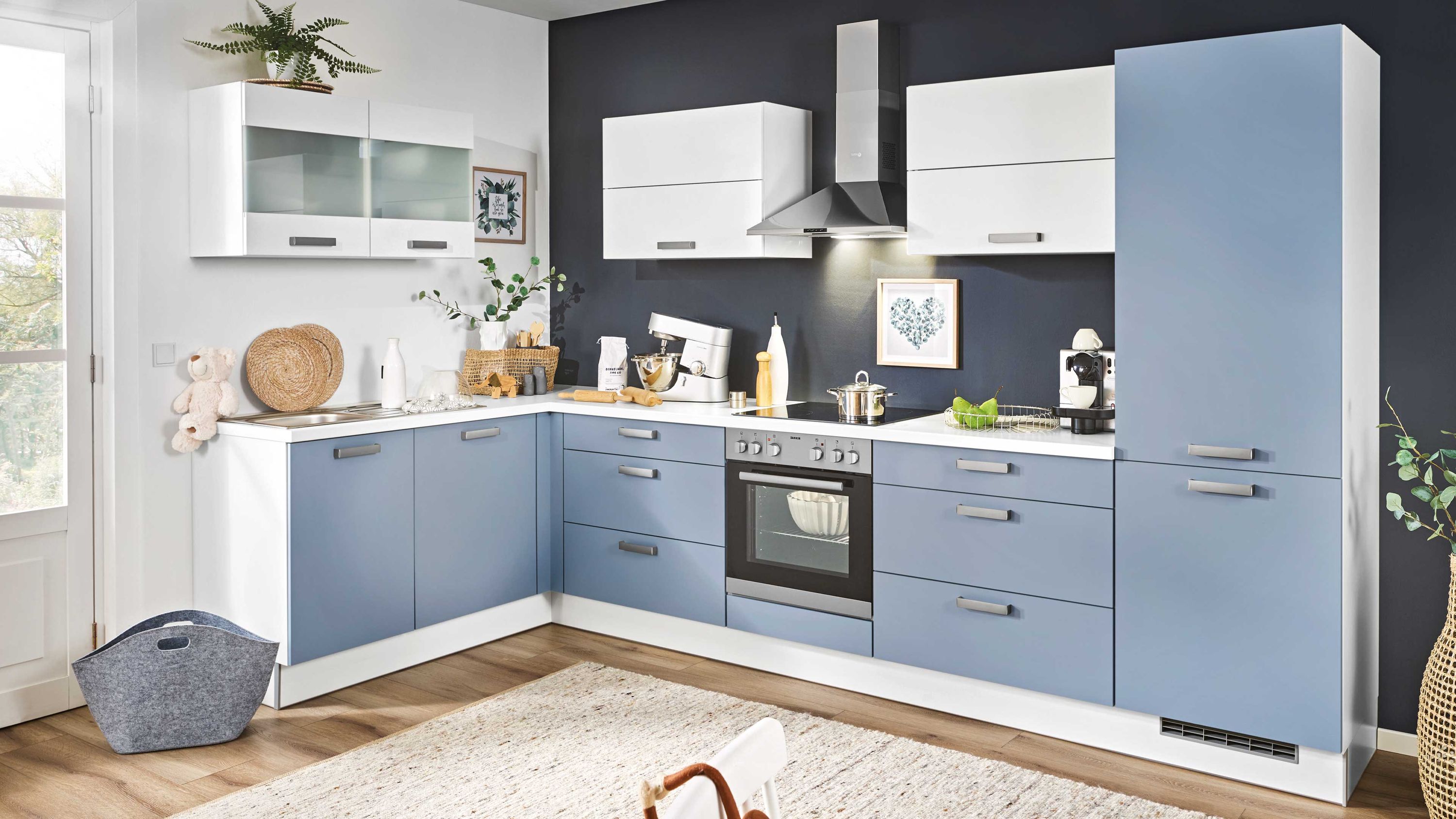 Eckküche mit blauen Fronten weißen Hängeschränken und Edelstahl-Dunstabzugshaube vor dunkler Wand.