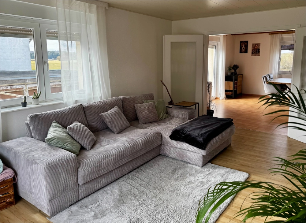 Modernes Wohnzimmer mit Blick ins Esszimmer Gemütliches Wohnzimmer mit grauem Ecksofa, Kissen, Teppich, Beistelltisch mit Lampe und großen Fenstern mit weißen Vorhängen.