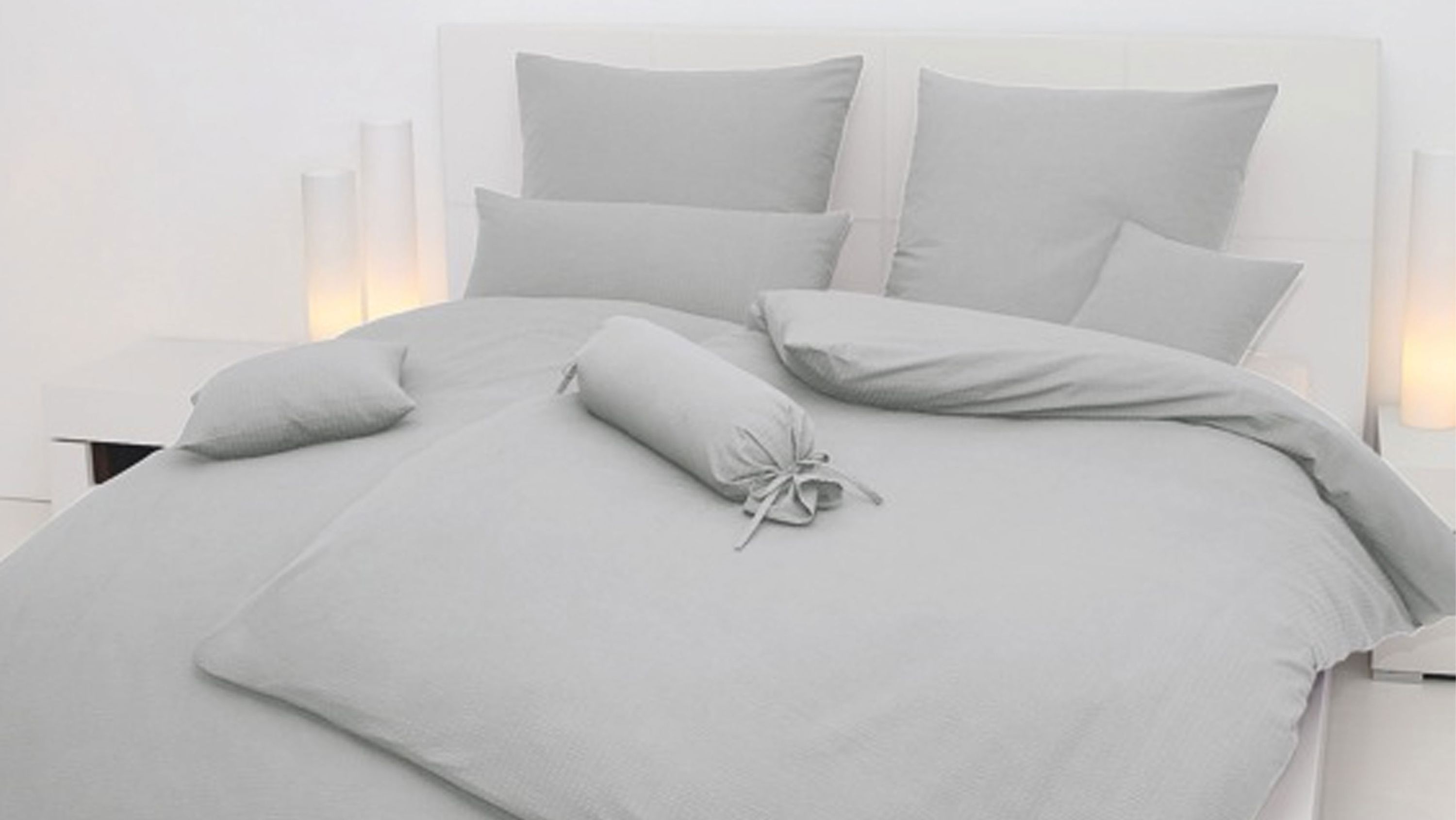 Hellgraue Bettwäsche mit mehreren Kissen und Zierkissen auf weißem Bett mit Kopfteil