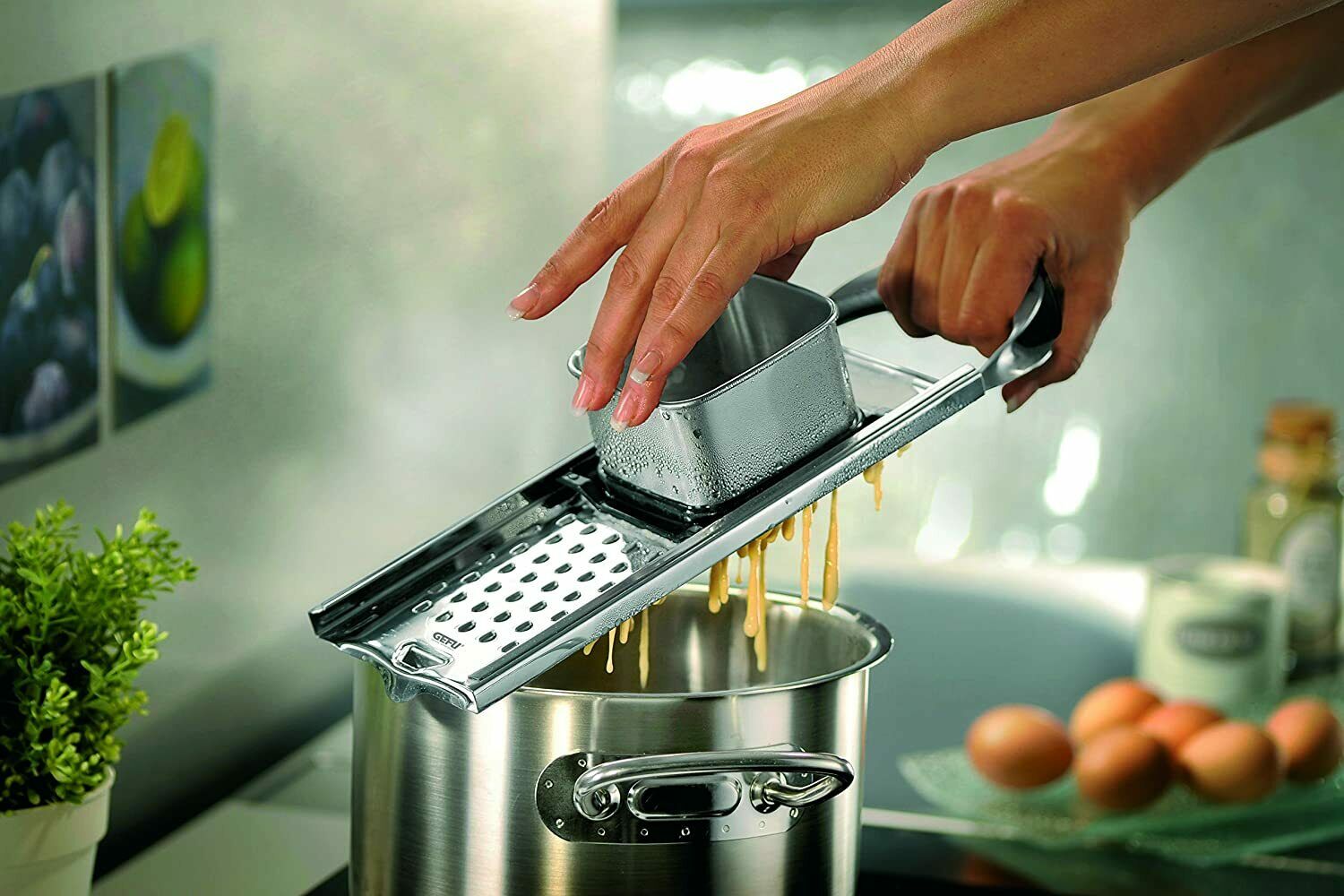 Person reibt Pasta mit Metallreibe über einem Topf in der Küche; im Hintergrund Eier und Küchenutensilien.