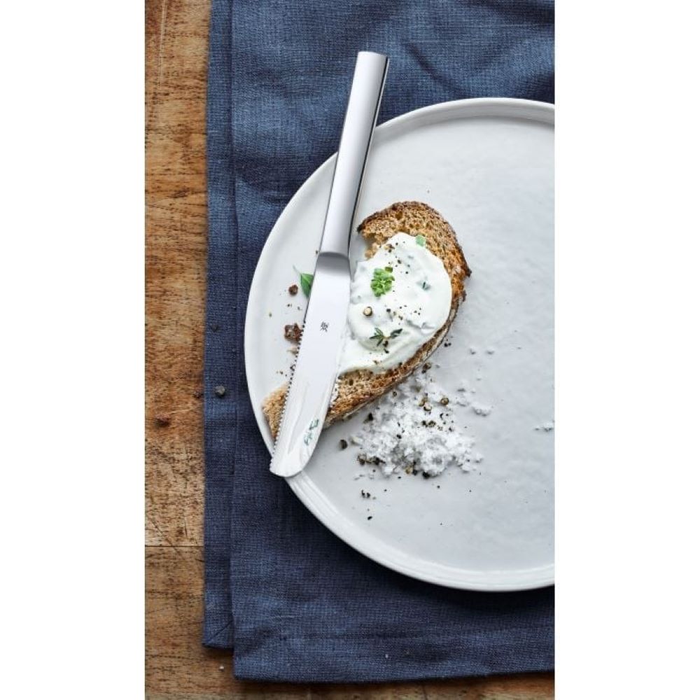 Edelstahl-Frühstücksmesser auf Brot – stilvolles Frühstücks-Setup mit blauer Serviette Frühstücksmesser aus Edelstahl auf belegter Brotscheibe auf weißem Teller dunkelblaue Serviette auf Holz frontal