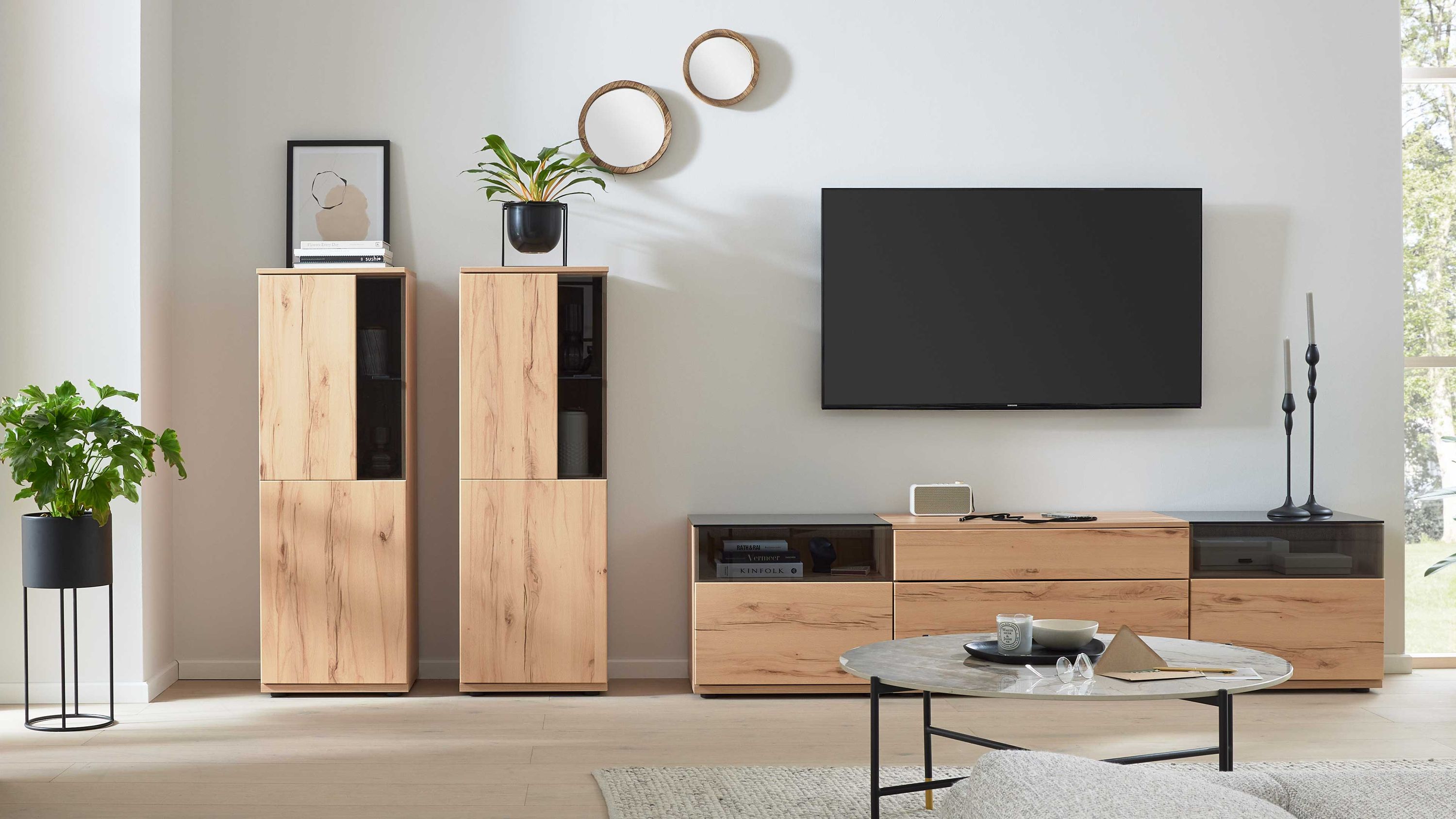 Wohnwand mit Schränken in Holzoptik und schwarzem Fernseher Wohnwand mit Schränken und Ablagen in Holzoptik schwarzem Glaseinsatz und Fernseher vor weißer Wand