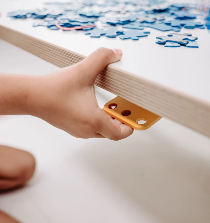 Poptop Schreibtisch Hand betätigt gelben Hebel eines höhenverstellbaren Schreibtischs mit blauen Puzzleteilen auf der weißen Tischplatte.