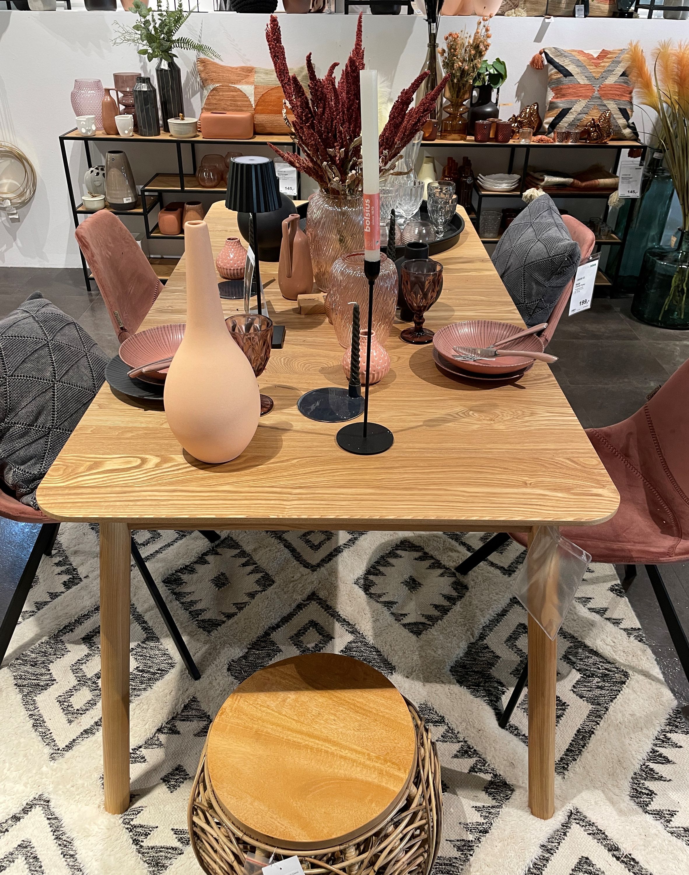 Modern gedeckter Holztisch mit rosa und schwarzen Dekoelementen, umgeben von gepolsterten Stühlen auf gemustertem Teppich.