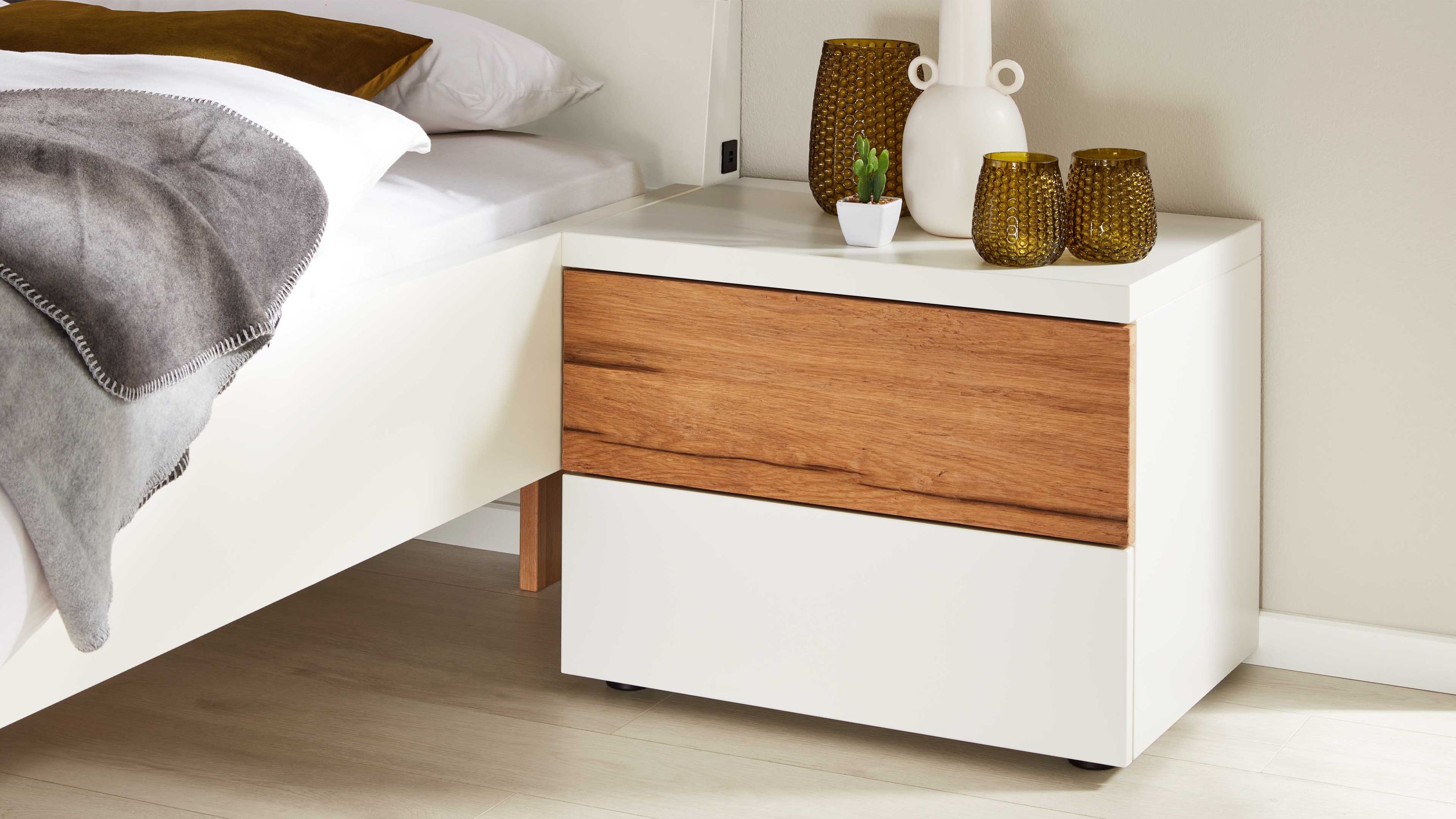 Weiße Nachtkommode mit Holzschublade neben Bett Weiße Nachtkommode mit zwei Schubladen obere Schublade in Holzoptik neben weißem Bett mit grauer Decke