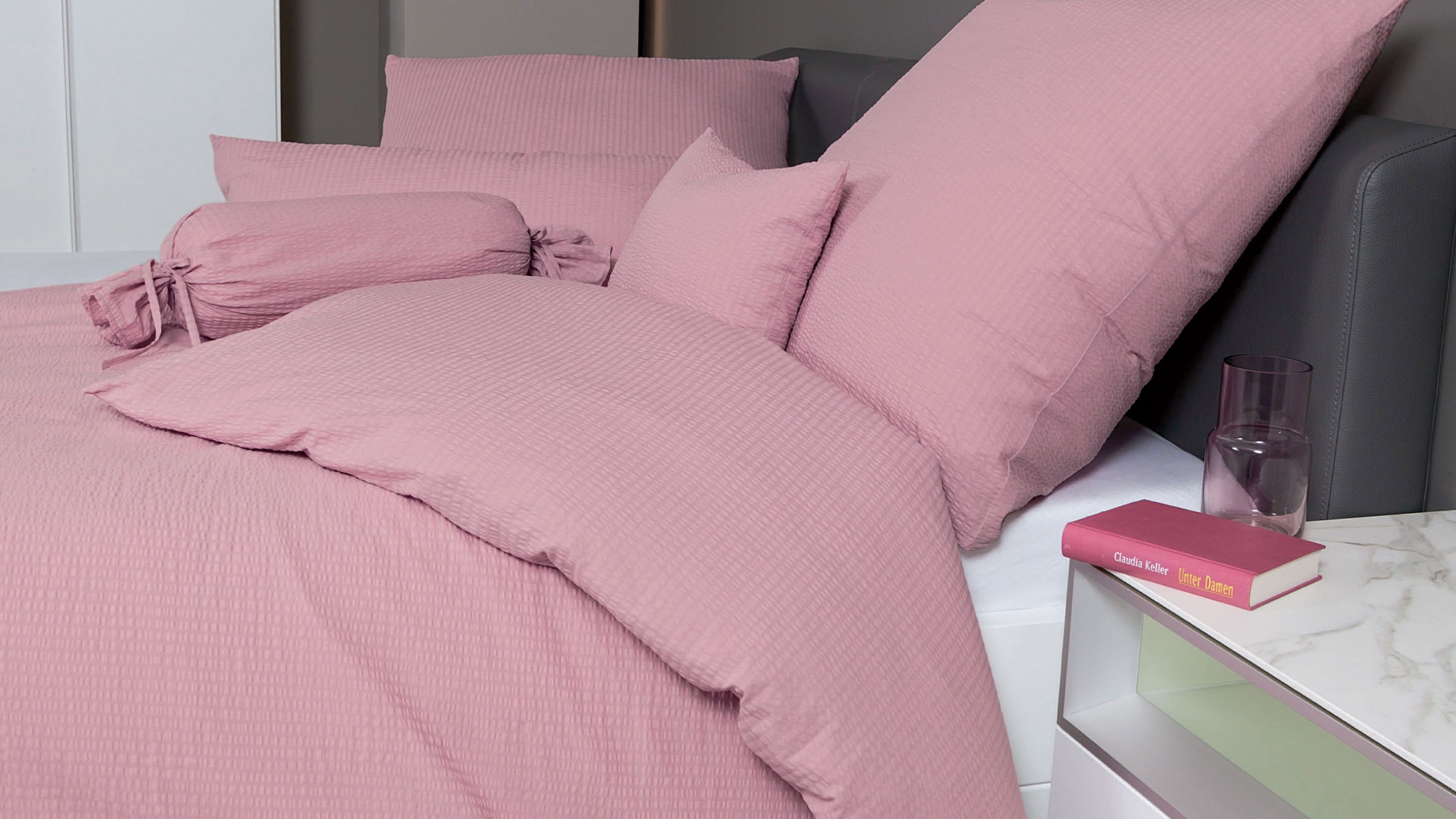 Rosa Bettwäsche-Set mit Kissen und Decke auf einem Bett mit grauem Kopfteil neben einem Nachttisch mit Buch und Glas