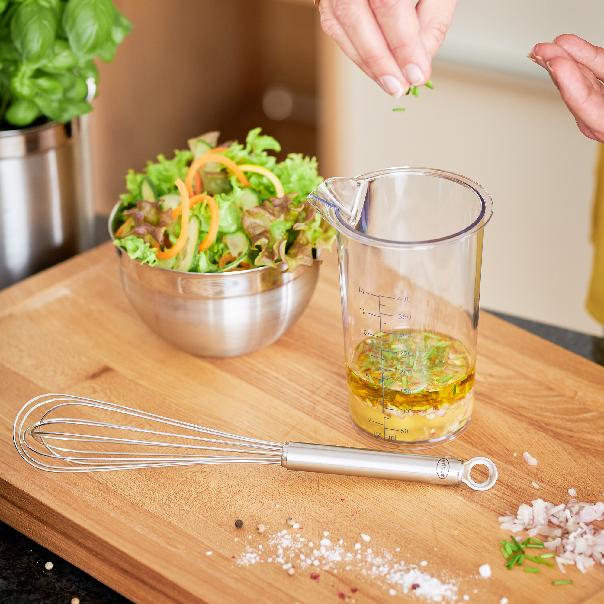 Edelstahlschüssel Silber Glänzende Edelstahlschüssel in Küchenszene mit Salat Schneebesen Zutaten und Öl-Messbecher auf Holzbrett schräg oben