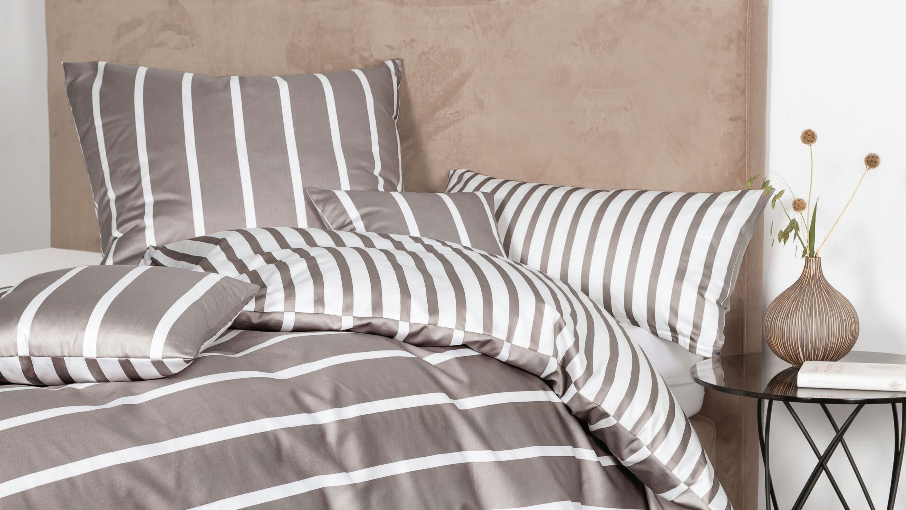 Bettwäsche-Set mit breiten weißen und grauen Streifen auf Kissen und Decke auf einem Bett mit beige gepolstertem Kopfteil