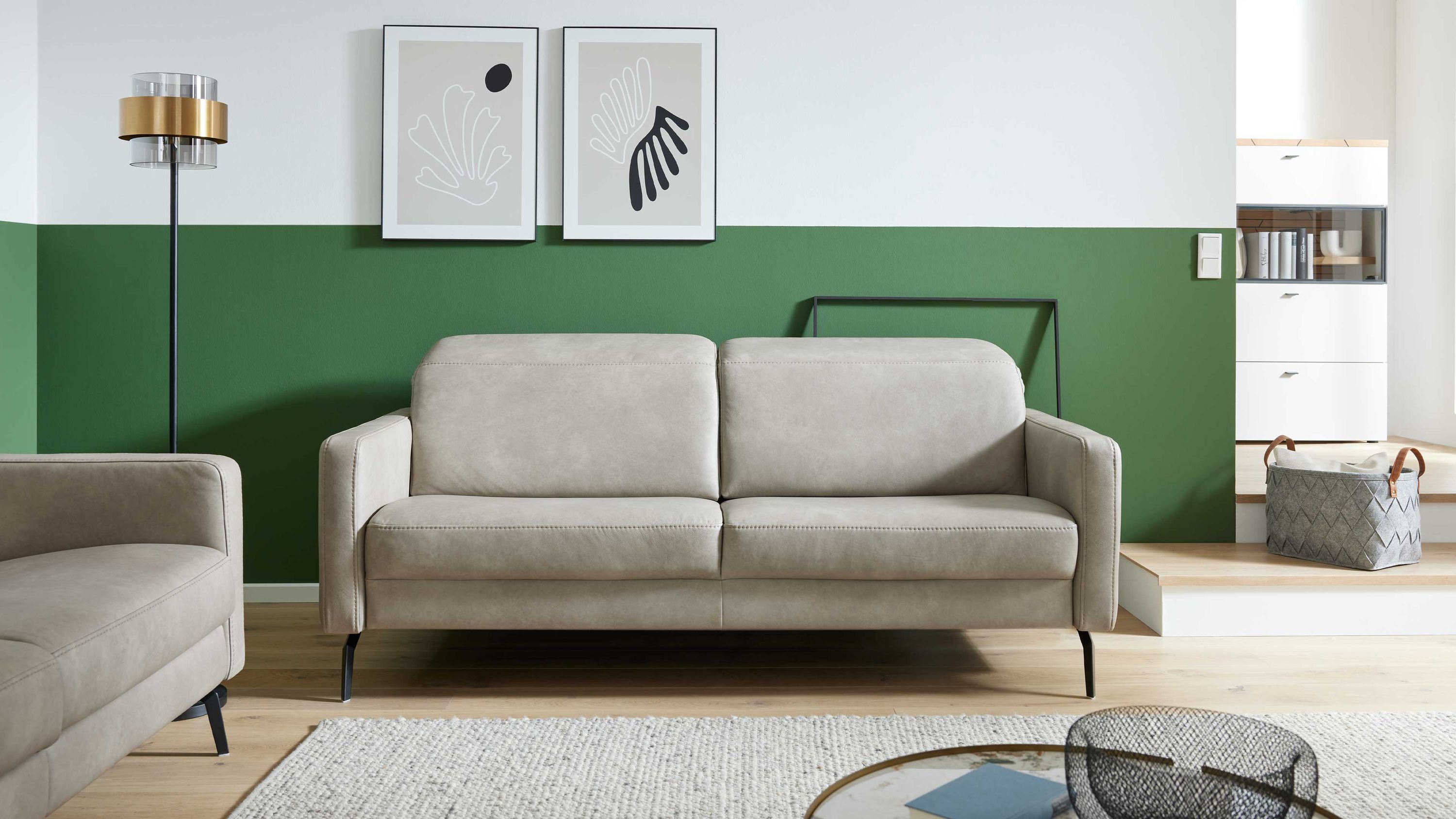 Zweisitziges Sofa in kieselfarbenem Leder mit schwarzen Metallfüßen vor grün-weißer Wand