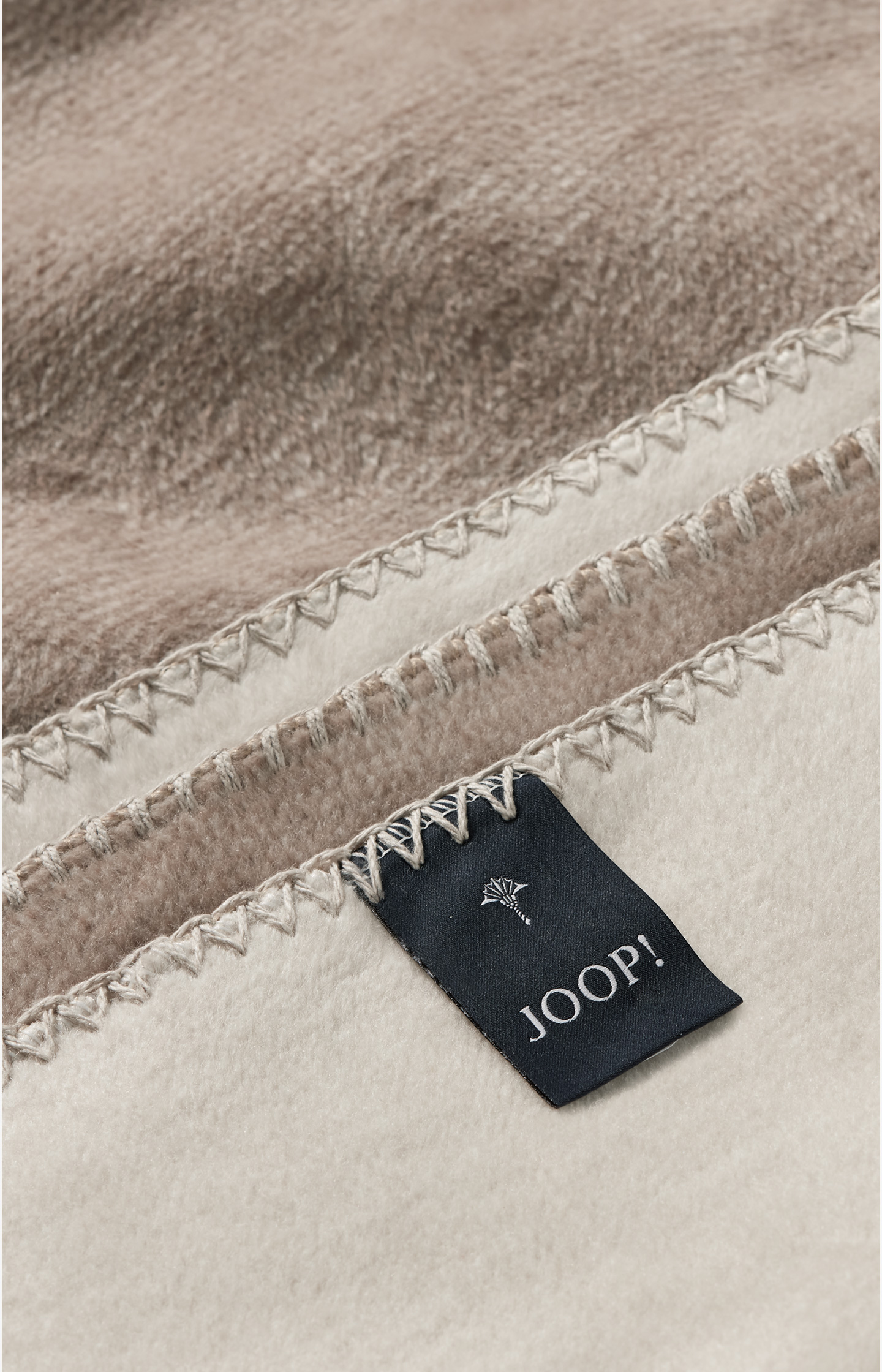 Nahaufnahme Kuscheldecke mit zwei Farbtönen in Beige und Sand sichtbare Kettelstichnaht schwarzes JOOP!-Label