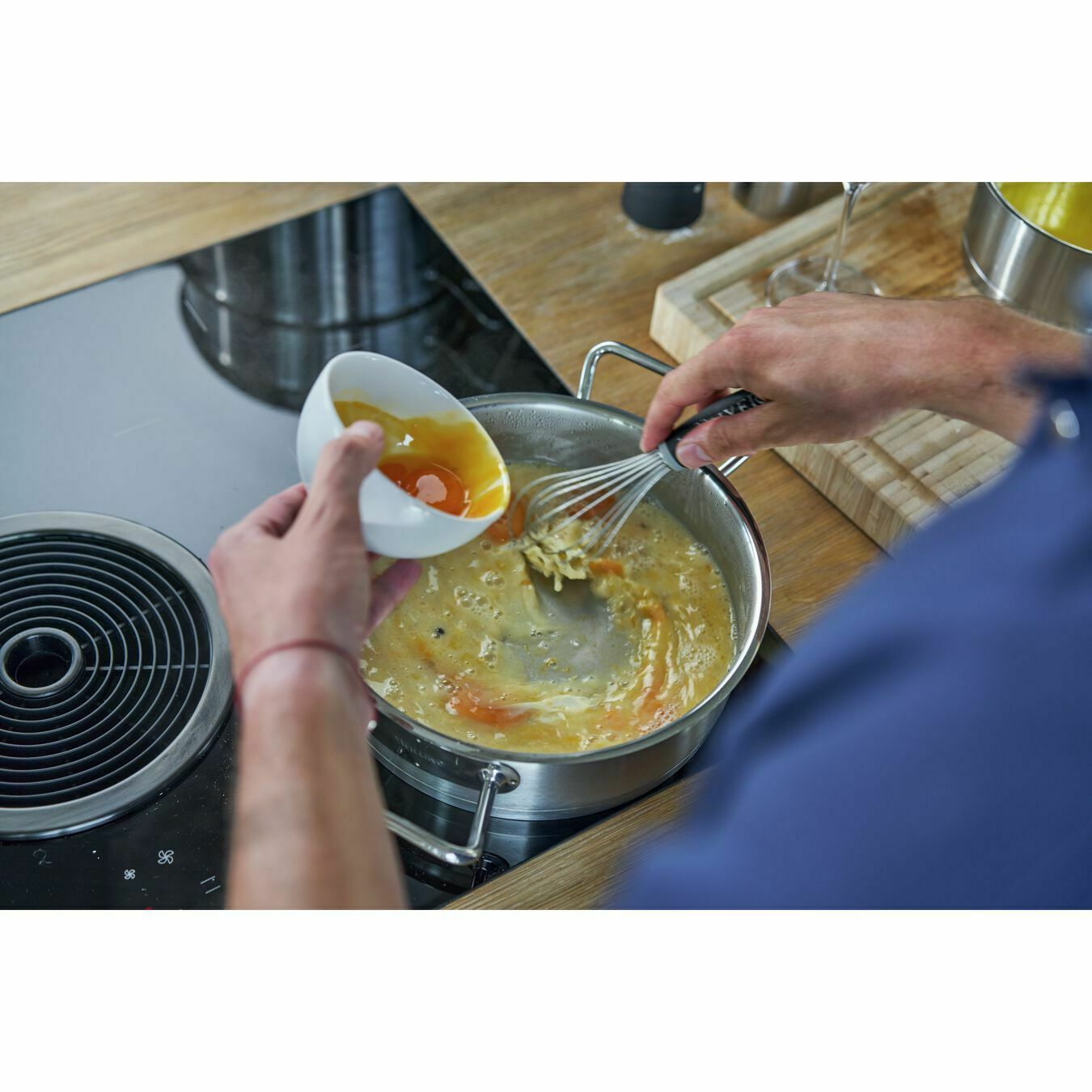 Person gibt Sauce in Pfanne auf Induktionsherd und rührt mit Schneebesen, schräg von oben aufgenommen