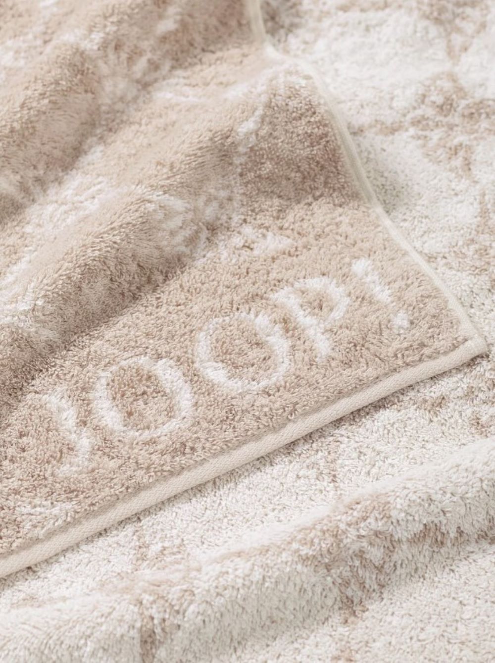 Beiges Duschhandtuch mit „JOOP!“-Schriftzug Beiges Duschhandtuch mit eingewebtem „JOOP!“-Logo in Großbuchstaben, von oben fotografiert.