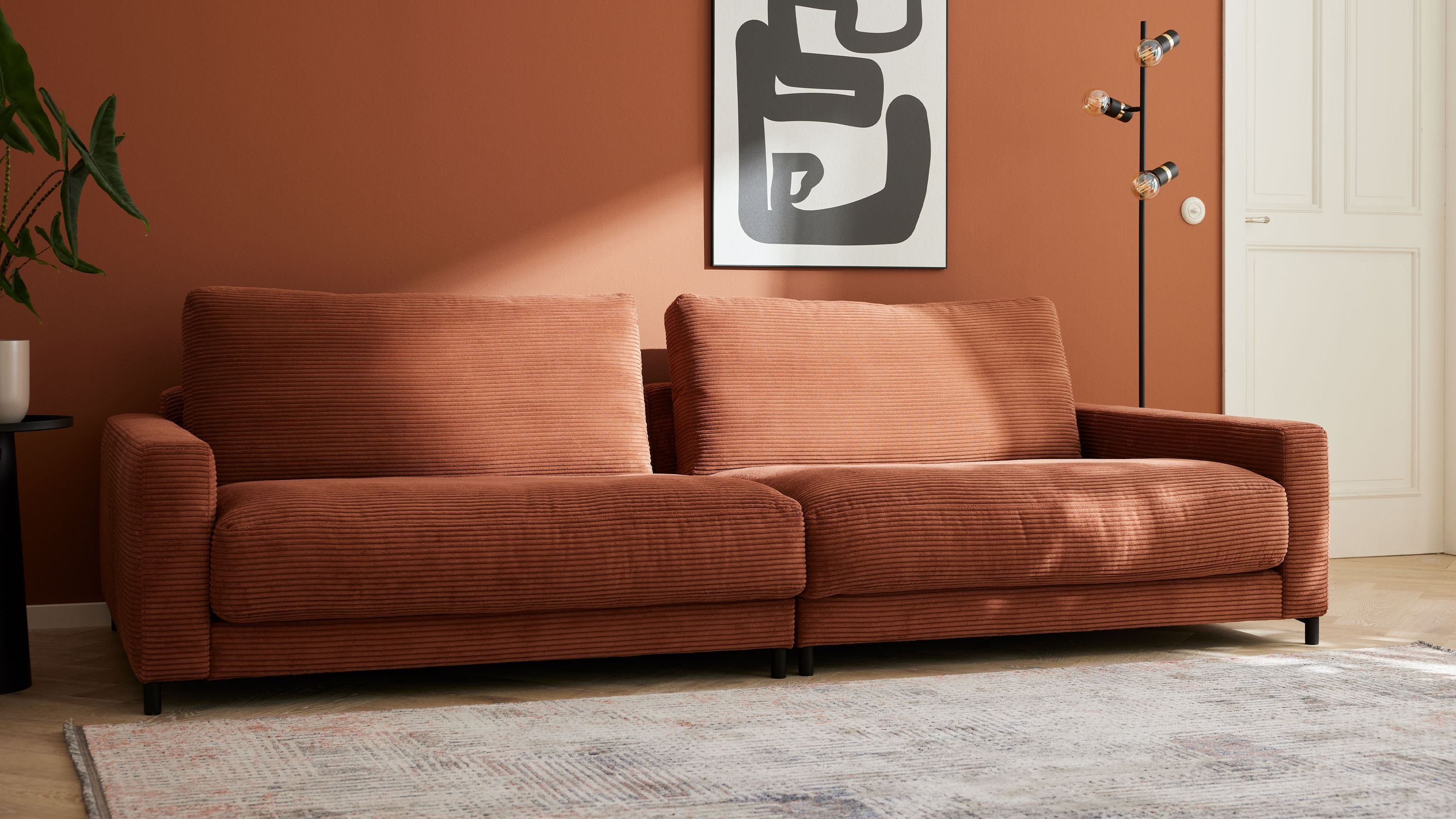 Großes Sofa mit rostfarbenem Cordbezug und schwarzen Metallfüßen vor rostfarbener Wand