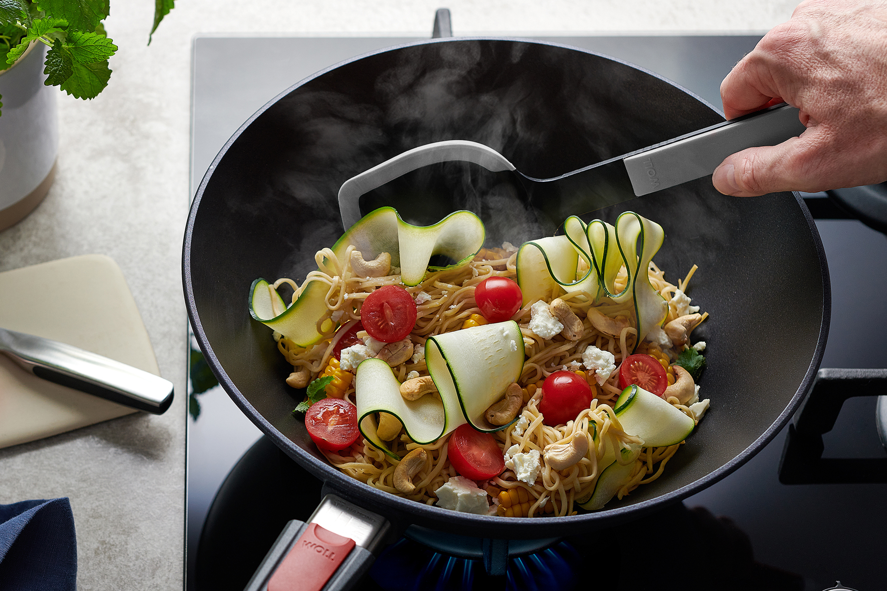 Wokpfanne von WOLL auf Gasherd mit dampfendem Nudelgericht, Zucchini, Tomaten, Cashews und Feta, Hand rührt mit Pfannenwender um.
