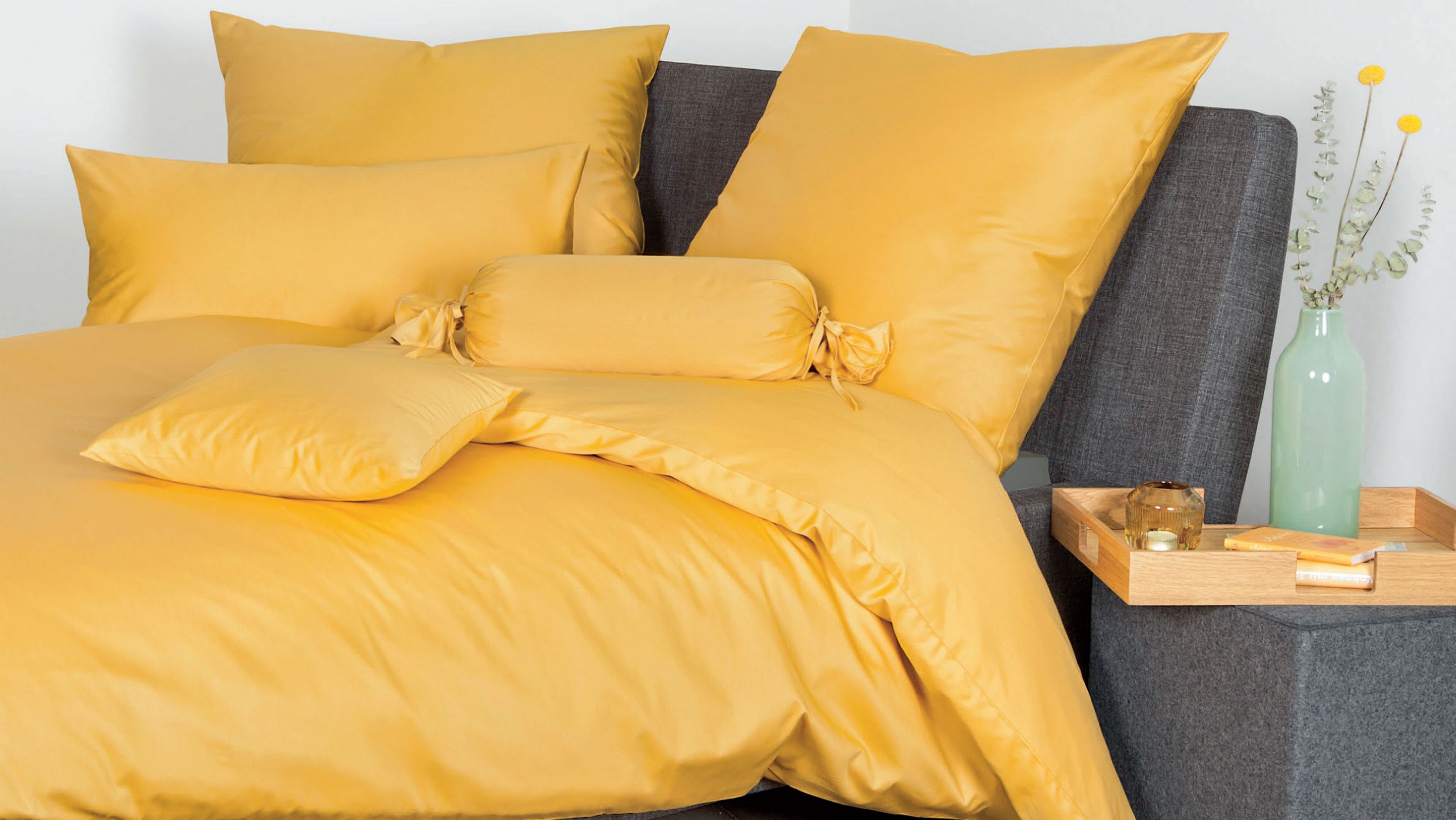 Gelbe Bettwäsche mit Kissen und Zierrolle auf grauem Polsterbett mit Holztisch und Vase