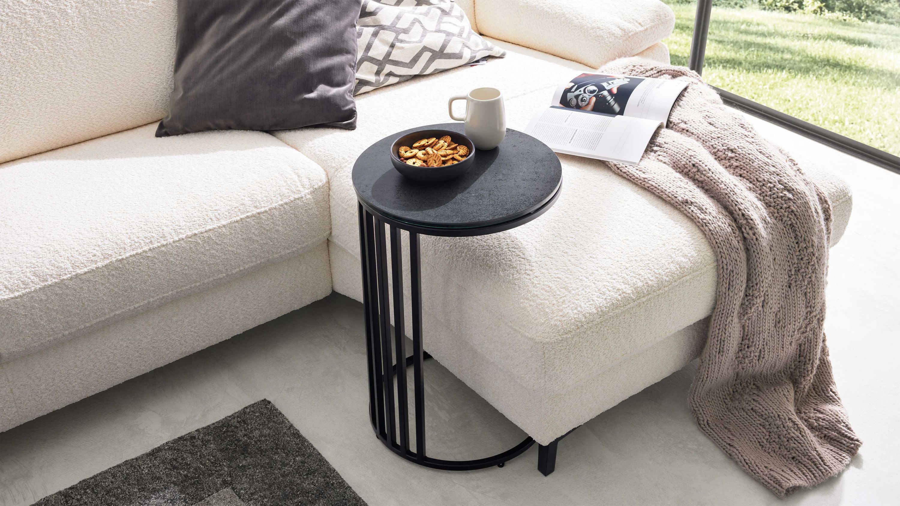 Runder schwarzer Beistelltisch neben Sofa Runder Beistelltisch mit schwarzer Tischplatte und schwarzem Gestell neben weißem Sofa mit Decke und Kissen