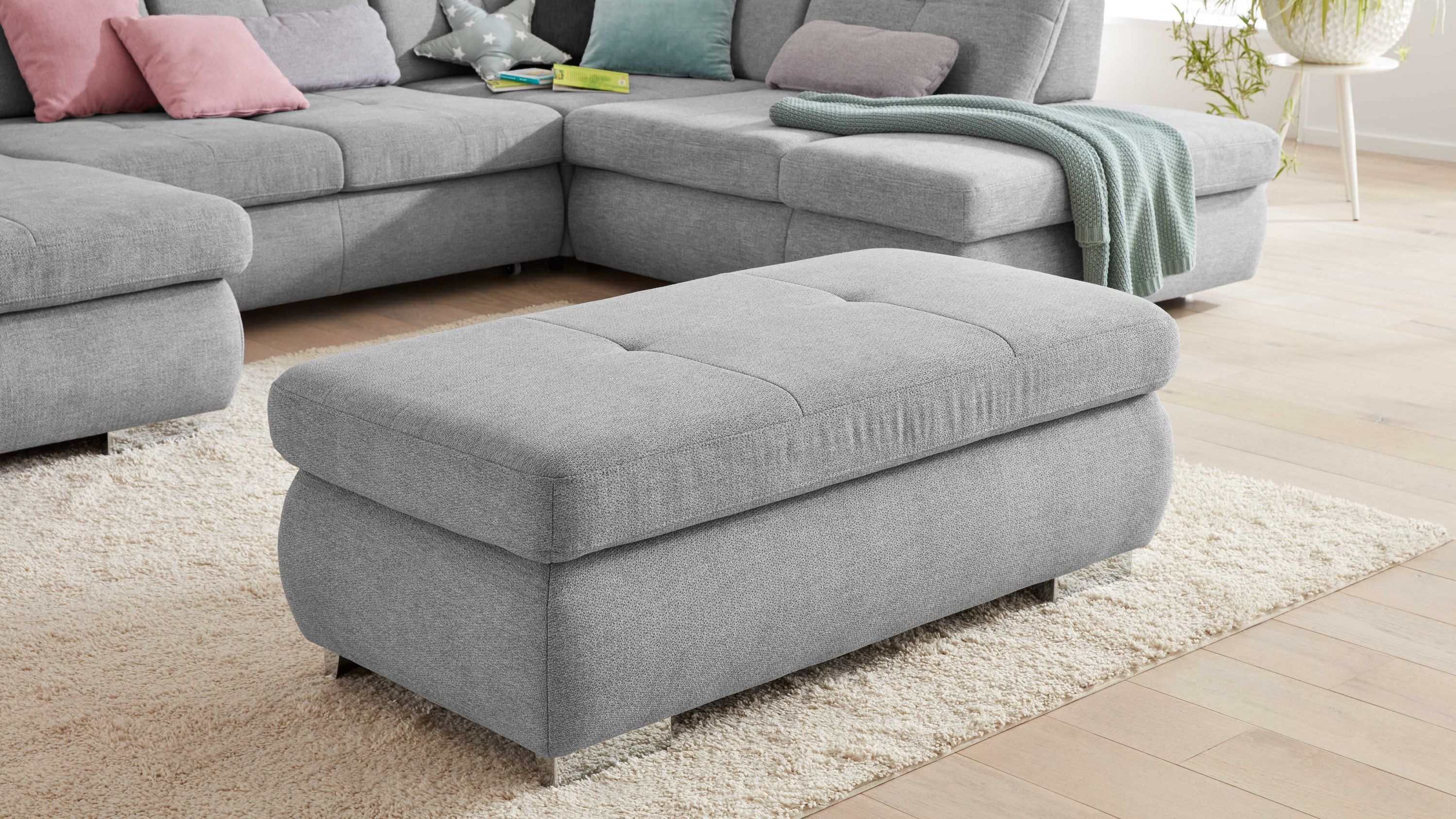 Rechteckiger grauer Hocker mit gepolstertem Sitz und Metallfüßen auf hellem Teppich vor grauem Ecksofa