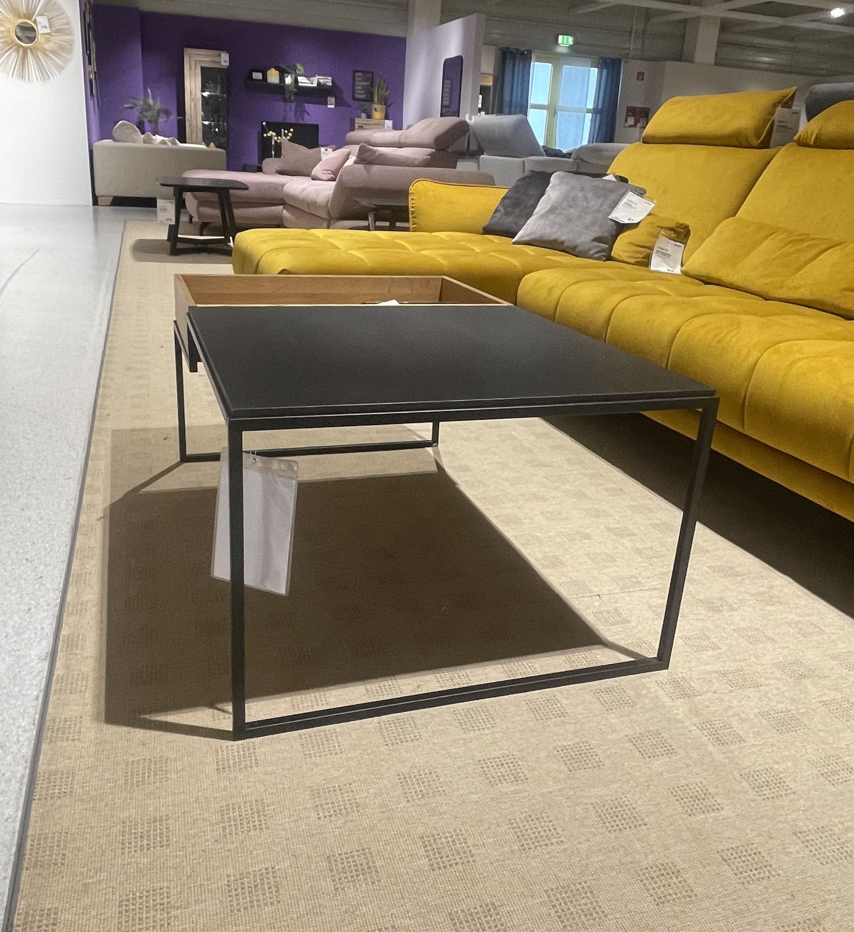 Moderner Couchtisch mit schwarzer Platte und schlichtem Metallgestell, seitlich angebrachtem Holzfach, vor gelbem Sofa platziert.